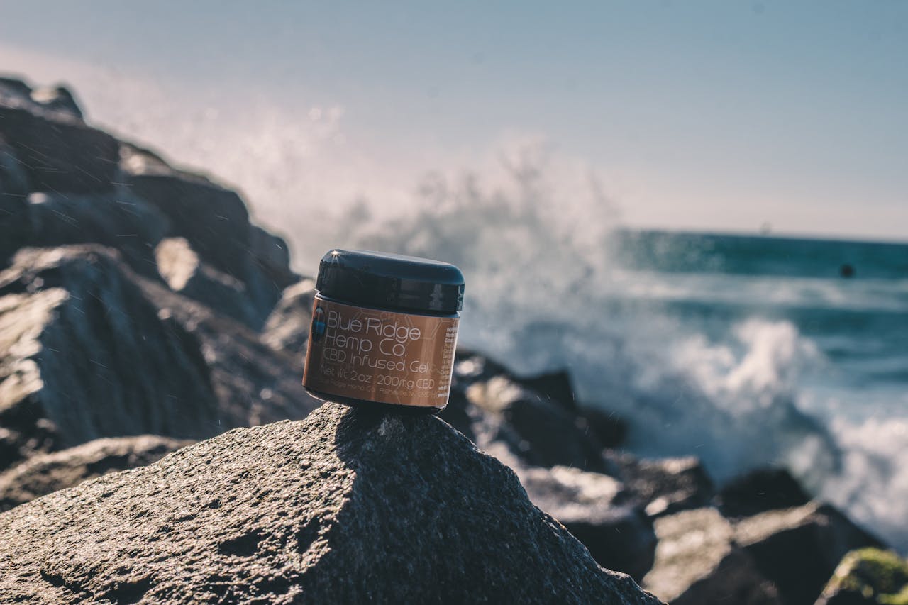 about-01 Close-up of CBD infused gel jar on rocks by the sea, showcasing product against a scenic ocean backdrop with splashing waves.